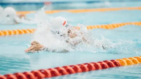 Hull City Council A swimmer with their arms outstretched and wearing a cap. The pool has red and yellow ropes marking the swimming lanes.