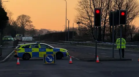 Danny Fullbrook/BBC A police car is parked blocking the A5. There are also cones and police tape restricting access. The sun is rising making the sky orange. More police vehicles are parked further along the road.