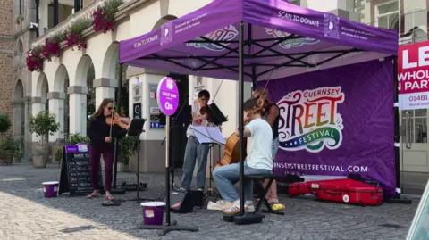 BBC A group of musicians playing instruments under a purple gazebo on the street in front of some shops. There are collection buckets on the floor in front of them. 
