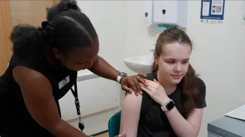 A woman giving a younger woman a vaccine in her arm. The older woman rests a hand on the other's shoulder and holds a needle in her other hand. The young woman looks ahead and has her left hand on her right shoulder. 