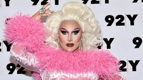 Getty Images The Vivienne pictured at an event in New York in 2022. She is in full drag, wearing a pink frilly top, and looking straight at the camera with her hand above her head