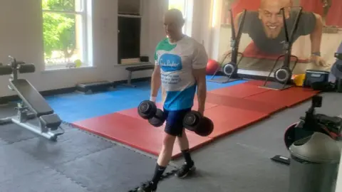 James Williams A man walking through a gym holding heavy weights. He is wearing blue shorts and white T-shirt with a Sue Ryder logo on it.