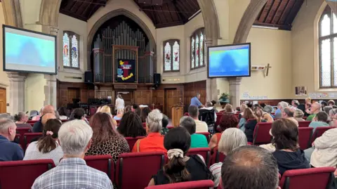 A church full of people sitting on rows of red chairs as they watch a memorial service. Two screens are mounted on the walls either side of the altar. A large organ is visible at the back of the room, in the middle of