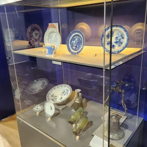 Scottish Maritime Museum A two-shelf glass display cabinet with four plates and a cup in blue and white on the top level, and a mix of items on the bottom including bowls, a small bell and a pair of small green glass bottles.