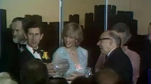 BBC specially shot Prince Charles is holding a crystal glass bowl and smiling at the crowd. He is wearing a black suit with a white shirt and a black bow tie. He has a daffodil in his lapel. Next to him smiling is Princess Diana, she is wearing a baby blue dress and a necklace. Her blonde hair frames her face and pearl earrings are visible. 