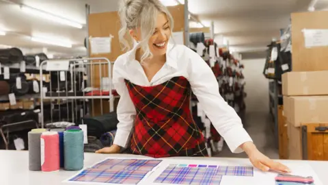 Designer Siobhan Mackenzie, a young blonde haired woman, leans over her white workbench where her tartan design is laid out, with samples of the thread colours. She wears a white shirt and a stylish tartan dress.