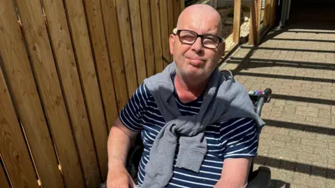 Helen Mahoney Bald man in glasses wearing striped shirt and grey jumper on shoulders sat in a wheelchair. 