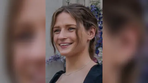 Family handout Close up of Olivia Wood's face, she has blue eyes and mouse blonde hair tied up. She is wearing a black top and stood in front of purple flowers.