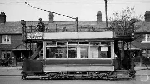 A black-and-white image of an old tram. The carriage has two levels, both lined with passenger seats. A conductor in a black hat is standing on the top floor. The tram runs along a cobbled street, past terraced houses. Text is just visible on an advert featured in a small panel on the top half of the carriage - it reads: "Bread and Fancies."