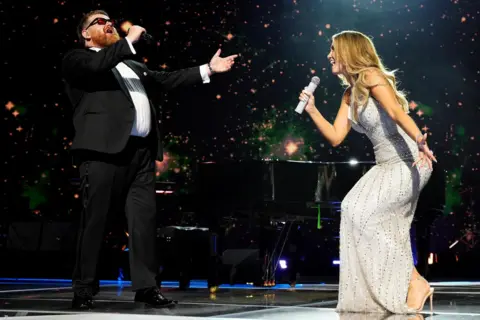 PA Media Cammy Barnes, wearing a black suit over a white shirt, sings with Delta Goodrem, who is wearing a long sparkly dress.