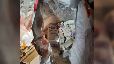 Large stacks of £20 notes bundled together with yellow and blue elastic bands. There are nine or ten bundles visible in the picture, though this is obscured as they are loose in a crumpled white plastic Tesco carrier bag. 