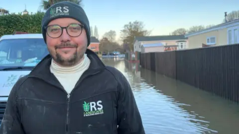 Man wearing glasses and woolly hat looking at the camera a van and flooding can bee seen behind him.