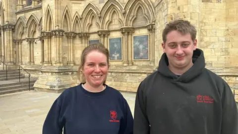 A young man and woman stand outside a historical building, wearing sweaters with the York Minster logo on the chest.