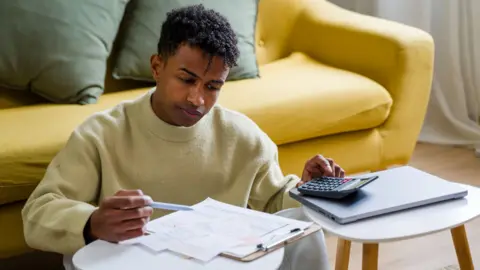 Stock photo shows a person looking down at a clipboad with bills and receipts while holding a pen and using a calculator in their living room at home.