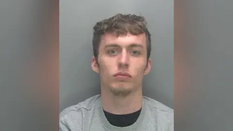 Cambridgeshire Police Custody photo of Callum Schooling against a plain grey background. He is wearing a crew neck grey sweatshirt over a crew neck black t-shirt. He looks directly at the camera and is not smiling. He has brown hair, blue eyes and facial hair on his chin.