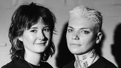 Zamaniego Studio A black and white headshot of two women. They are both smiling into the camera. The woman on the right has tattoos on her neck as well as short, light hair and the woman on the left has a nose piercing and feathered dark hair.