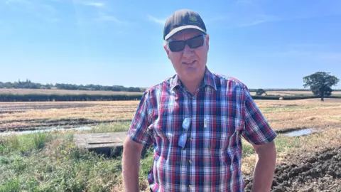Kate Bradbrook/BBC Peter Howard is standing in a partially flooded field. He is wearing sunglasses, a peaked cap and a checked shirt.