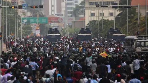 Getty Images Thousands of Sri Lankans gather in a road. Above them are road signs and traffic lights and in the centre of the picture are three large police vehicles, amidst the crowd. 