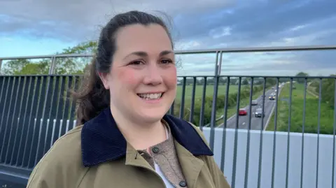 Tom Jackson/BBC Fiona Reardon-Rose standing on a footbridge with high railings behind her. She has dark brown hair pulled back from her face and is wearing a tan jacket with a blue cord collar. Behind her and through the railings can be seen the single carriageway A10, with cars travelling in both directions and grass verge, hedgerows and fields on either side.