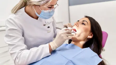 Getty Images A woman lying on a dentist's chair with her mouth open - a dentist is examining her teeth