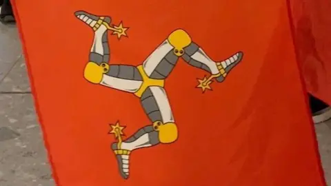 BBC A close-up of three Manx flags, which are red with a yellow, white and grey three-legs of man symbol. 