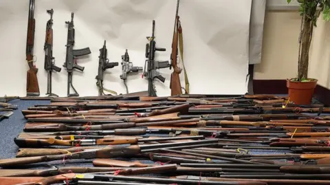 IOM CONSTABULARY A room full of firearms, they are all long wooden firearms, some are lying on the floor others are resting against the wall.
