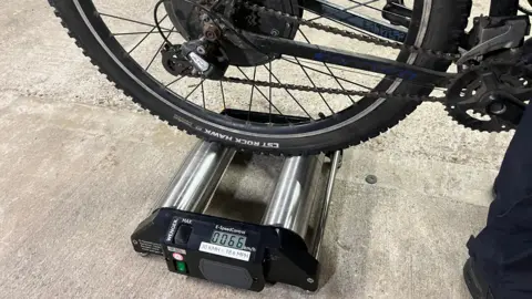 O roată de bicicletă neagră se află deasupra unui dispozitiv metalic mare, care are doi cilindri argintii și un indicator numeric în partea de jos.