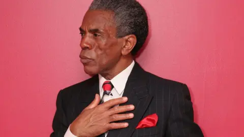 Lia Chang André De Shields, dengan garis-garis rambut beruban, memegangi jaket dan dasinya yang bercorak arang. Saputangan merah menyembul dari sakunya. Dia digambarkan di dinding merah.