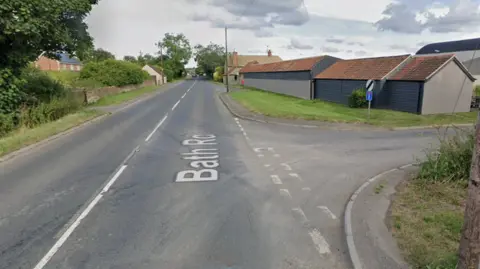 Google Maps The junction at the A365 and Bowerhill lane. There are farm-like buildings to the right of the carriageway and a grassy verge either side.