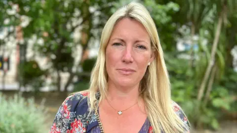 NIKKI FOX/BBC Emma Cracknell, daughter of Susan Spring standing in the gardens opposite Arundel House in central london. She is looking directly at the camera and has long blonde hair. She is wearing a navy floral v-neck top with a small cross on her necklace.
