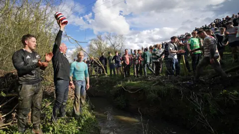 PA Media A man holding a barrel aloft by a stream, with the other side of the bank filled with people congratulating each other