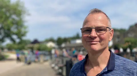Heidi Tomlinson/BBC A man with short grey hair and glasses smiles into the camera. He is wearing a navy polo shirt. The background is blurred, but it is a public place on a sunny day.