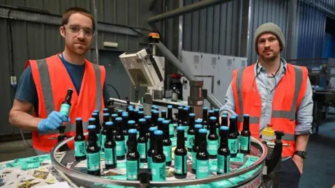 Getty Images Dickie and Watt wearing high‑visibility vests stand beside a circular bottling table filled with BrewDog beer bottles inside an industrial brewery, with production equipment visible in the background.