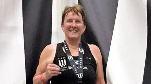 Carole Munro smiles at the camera wearing a medal and holding a patch. She is standing in front of a black and white striped backdrop and is wearing a black vest top.