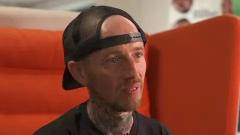 Steven, a man in a black t shirt and black snapback cap sat on a large orange chair. He has a short red haired goatee.