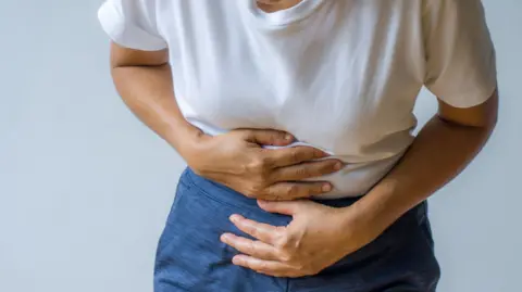 Getty Images A person is holding their tummy with both hands. They have a white t-shirt and navy trousers on. The background is white. 