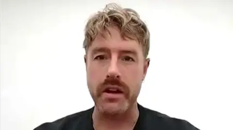 Christian Aldridge sits in front of a white wall while talking. He has blonde hair and a trimmed beard. He is wearing a black top.