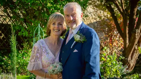 Kate Turner Kate and Barry on their wedding day in a mid shot smiling to camera. Kate is wearing a pale pink floral lacy dress with capped sleeves and is holding a small posy of flowers. Barry is wearing a dark blue three piece suit with a spray of flowers in his button hole. There is greenery behind