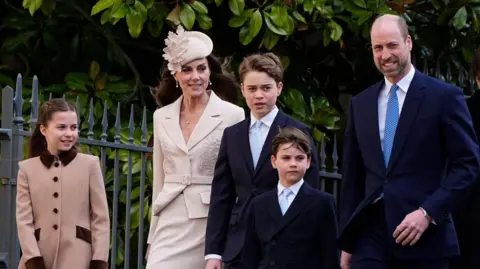 Prince of Wales, William, Princess of Wales, Catherine and their children - George, Charlotte and Louis walk. William, George and Louis are wearing blue suits and ties. Catherine is dressed in a beige skirt and jacket and Charlotte in a beige coat. 