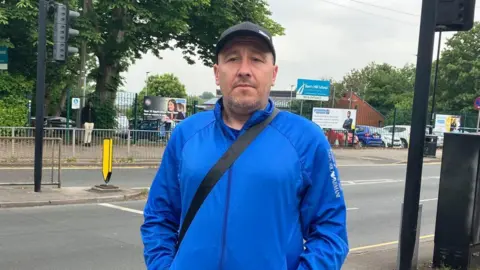 A man on a pavement in front of a road. There is a school in the background. He has his hands in his pockets and is wearing a blue jacket. He is wearing a black cap and a black messenger bag.