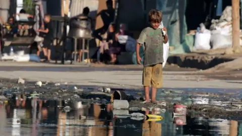 Getty Images A child in dirty clothing stands in dirty water and rubbish with a plastic bag on his shoulder in Gaza