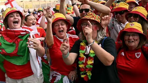 A large group of Wales football fans, with mostly women in the front, dressed in red football shirts and colourful bucket hats, mouths open in chants with their arms in the air.
