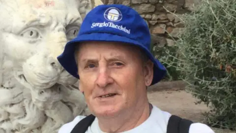 Stanley Smith smiling and wearing a blue bucket hat with the words Sergio Tacchini written on in white lettering. He appears as though he is on holiday in a park. A stone lion is behind him. He has a small stud earring and wears a white T-shirt. The straps of a black backpack can also be seen.