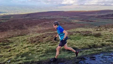 David Griffin David Griffin is running along a moorland. He is wearing a blue shirt, with a lighter blue vest and gloves. It is very muddy with a grey blue sky.