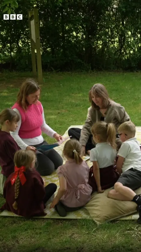 A group of children and two adults sitting on a blanket in a green space, surrounded by trees.