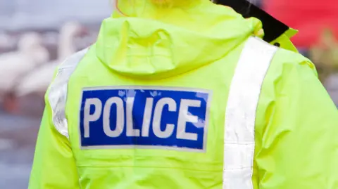 A female police officer from behind. Her jacket is bright green and has "Police" written in white font in a blue box