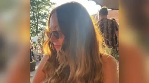 Family photograph Stephanie Blundell, 41, poses wearing glasses while at an outdoor event. She has long brown hair and is smiling while looking to her right.
