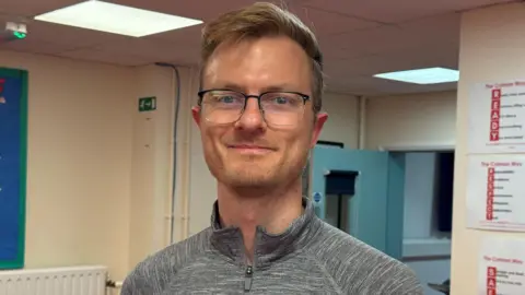 Cameron Noble/BBC Teacher Michael Burdett stands in a school corridor and wears spectacles