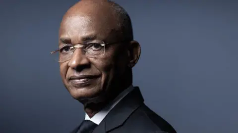 AFP via Getty Images Guinean economist and politician and former Prime Minister of Guinea Cellou Dalein Diallo wearing a dark formal suit, stands against a smooth, muted blue-grey background. He is positioned slightly to the side, with part of the upper body and shoulders visible.