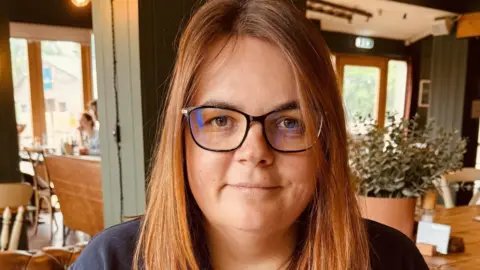 Alderford Lake A woman with brown hair and glasses, pictured from the shoulders up, looks at the camera. There is a cafe/restaurant interior in the background with tables and chairs and large windows.